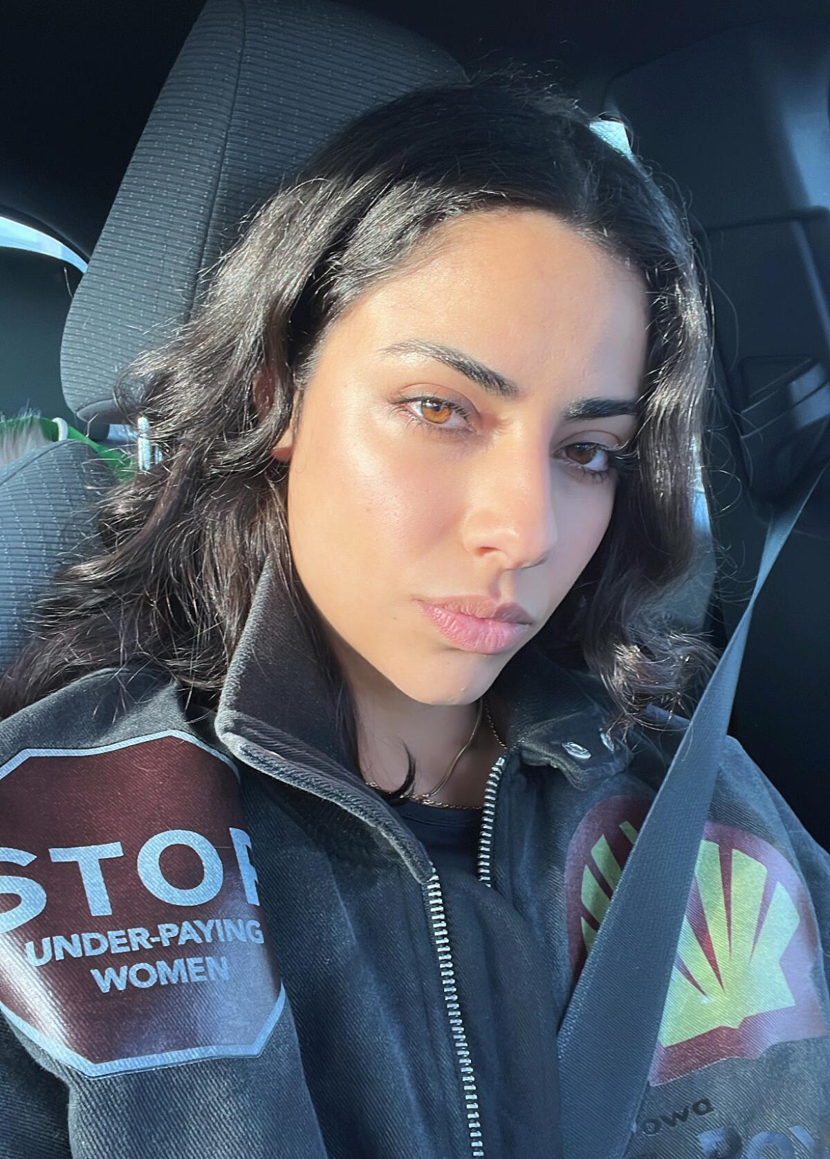A photo of a woman with dark hair in a jacket with a patch of a red Stop sign and another of a yellow shell outlined in red