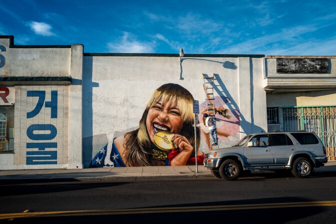 A man on a ladder paints on a white wall. In the painting a woman with brown and blonde hair smiles while taking a bite of her gold Olympic medal. Portions of the American flag can be seen wrapped around her shoulders.