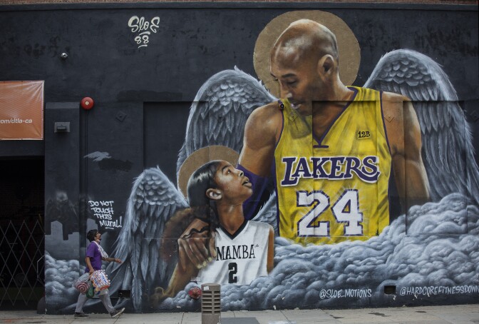 A tall man wears a Lakers jersey. He has his arm around a small girl who has a white basketball jersey that reads, "MAMBA." The two have angels wings behind them.
