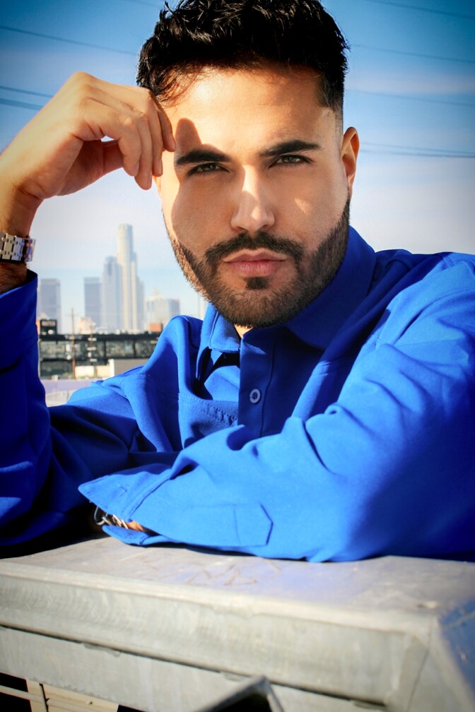 A younger man with fair skin, dark hair and a beard wears a blue shirt and poses for the camera. The downtown L.A. skyline is visible behind him.