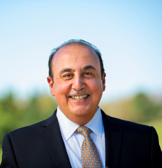 A man with medium skin tone and a gray mustache smiles at the camera. He wears a blue suit and a patterned tie.