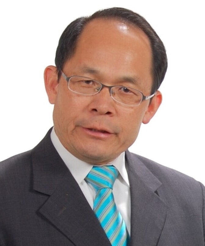 A man with medium skin tone, dark hair and a receding hairline looks at the camera. He wears a suit and a teal striped tie.