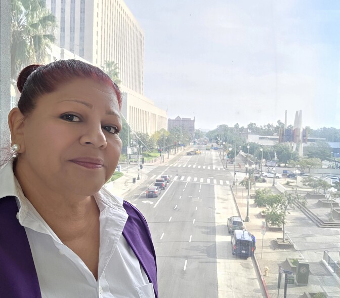A woman with medium skin tone and reddish brown hair wears a purple jacket and white shirt. A city street is visible in the background.