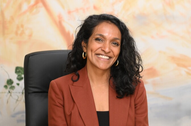 A woman with medium skin tone and dark hair smiles at the camera. She wears a brownish blazer and black shirt.