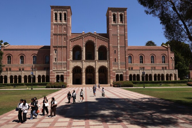 People walk in a large plaza in front of a large brick collegiate building. Lawns flank the plaza, which is partially shaded by a tree.