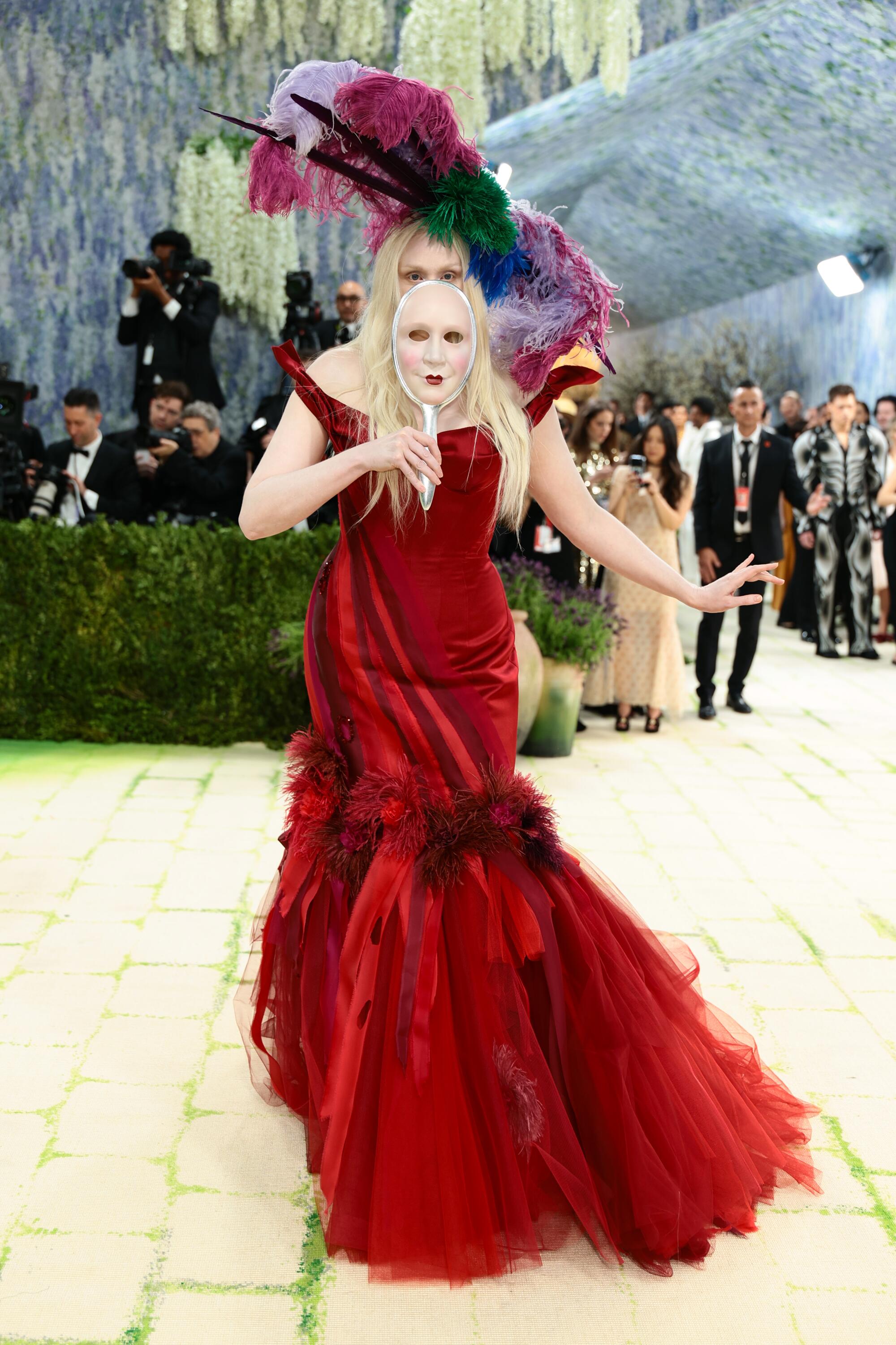 Gwendoline Christie, wearing a red gown and holding a mask, poses at the Met Gala. 