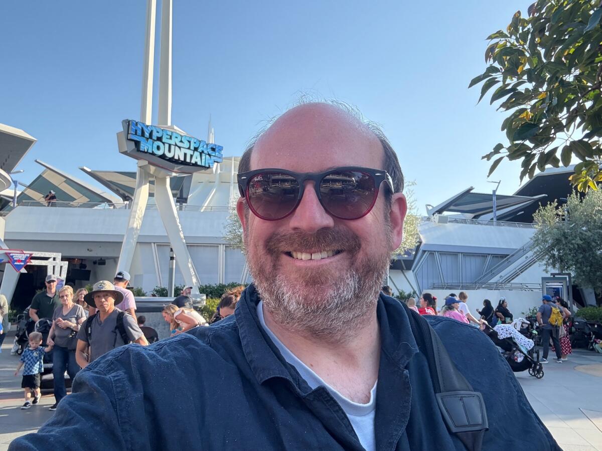 A bearded man in sunglasses in front of a theme park ride. 