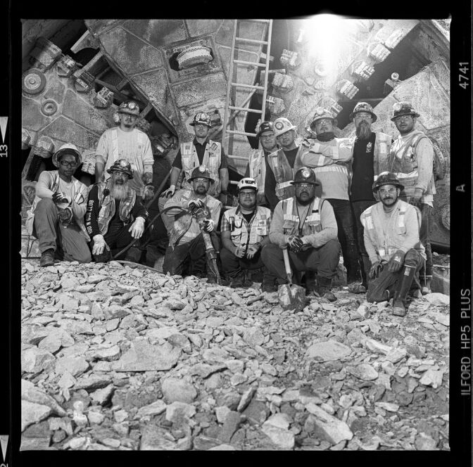 A group of thirteen people in construction clothes, including helmets and high-viz vests, stand in two rows looking at the camera. Some have tools, like shovels. The image is black and white. In the foreground in front of the workers, is a collection of broken rocks. There is a ladder in the background behind the workers. They stand in front of a tunnel boring machine.