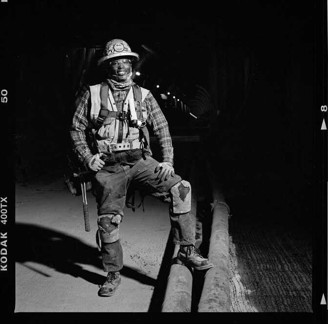 A black-and-white photograph of a woman in a construction helmet and vest. The woman is smiling and looking directly at the camera. She's wearing a plaid shirt and dark jeans. Her left hand rests on her left leg, which is propped up. Other than a few small lights, the background of the image is mostly black.