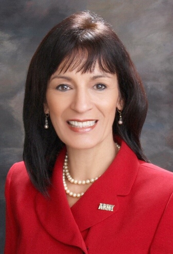 a smiling woman with shoulder length brown hair, a pearl necklace, and a pin that says "ARMY" pinned to the lapel of a red coat