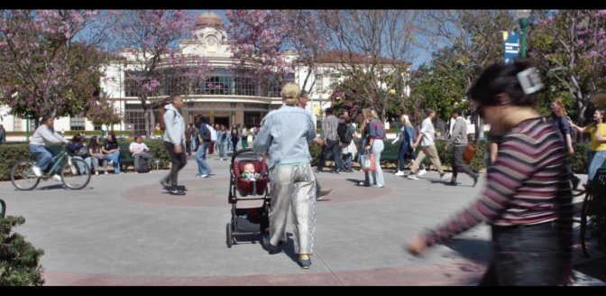 A cement and brick campus quad with students walking around, one in the center only visible from behind, headed toward a domed building and pushing a baby in a stroller. Trees with purple flowers are in front of the building.