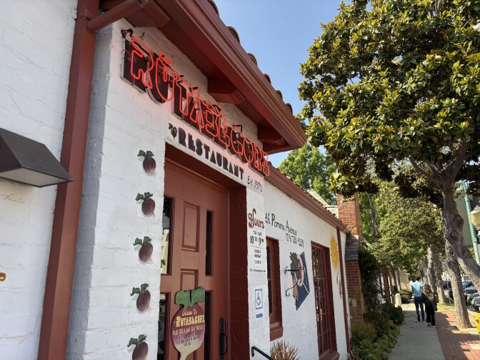 The outside of a one-story white brick building with a Spanish tile roof on a tree lined street. The sign about the door reads "Rutabegorz" in red neon, with cartoon rutabegas painted along the left side of the door.