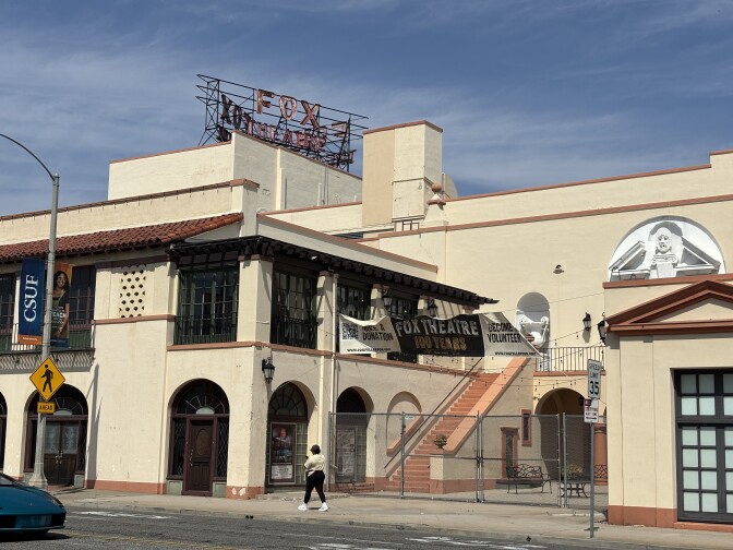 An across the street shot of a two-story tan Spanish style building with an old neon sign on a wire structure that reads "Fox Theatre" on its roof. A chain link fence blocks the first-floor entrance.