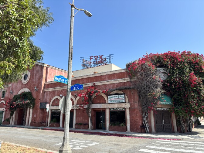 A sun bleached red building with tan arches and a sign that reads "Angelo's and Vinci's. Bougainveilla vines with red flowers cover the corner entrance of the building. A red sign that reads "Fox Theatre" is visible atop the roof of a tan building in the background.