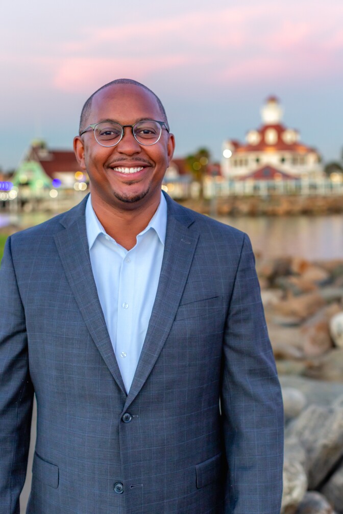 a smiling man with a shaved head and glasses wearing a blue suit jacket over a light blue collared button up shirt