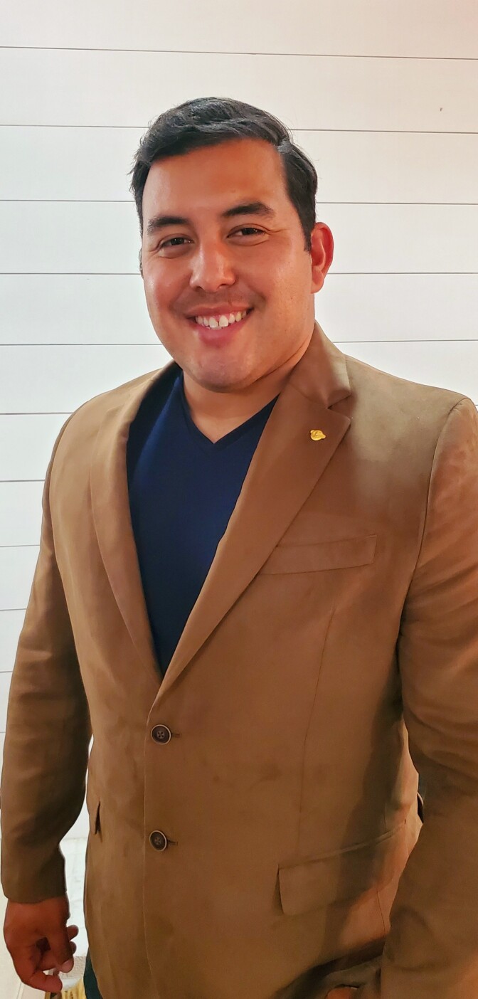 a smiling man with short dark hair wears a brown blazer over a dark blue v-neck shirt