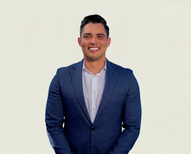 a smiling man with short dark hair wearing a dark blue suit jacket over a light blue collared button up shirt