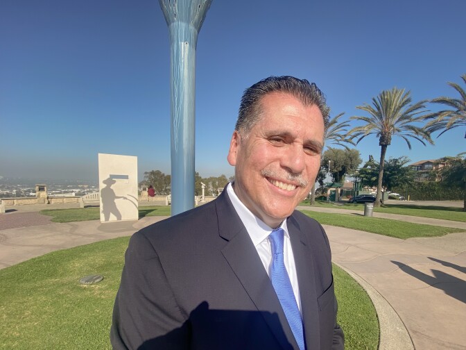 A man in a dark suit and blue tie smiles for the camera. Behind him we see a park and some sort of public monument.