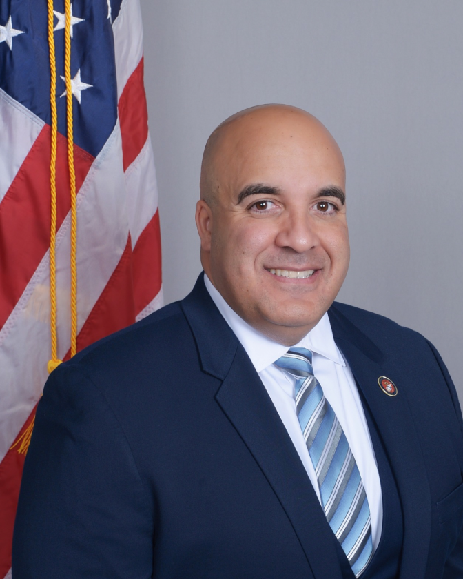a smiling man with a shaved head wears a blue suit jack and blue striped tie with a white collard shirt and stands in front of an American flag