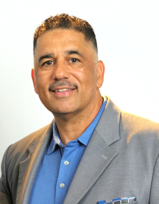 a smiling man with short dark hair wears a gray suit jacket over a blue button up collared shirt