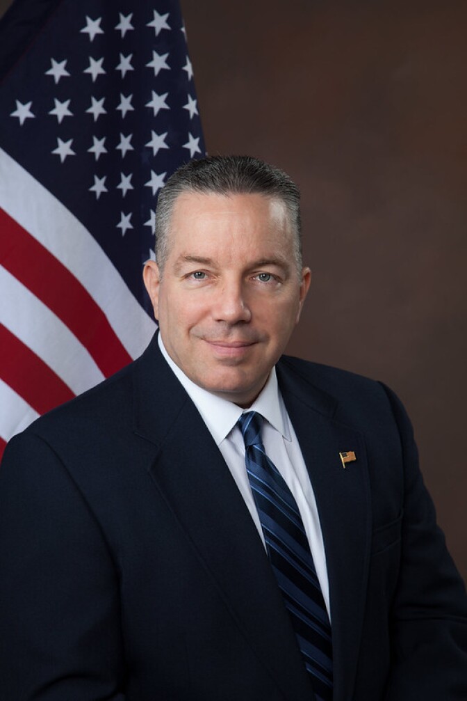a smiling man with short graying hair with a dark suit jacket and blue striped tie with an American flag pin on his lapel sits in front of an American flag