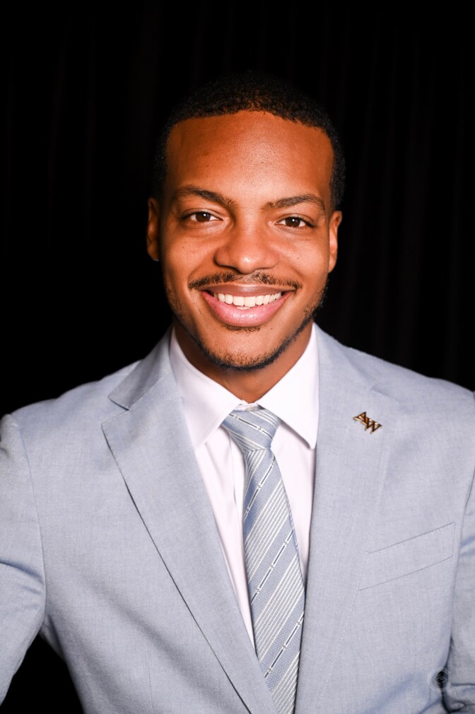 a smiling man with short dark hair wears a light blue-gray suit jacket and a light blue-gray striped tie with a white button up collared shirt