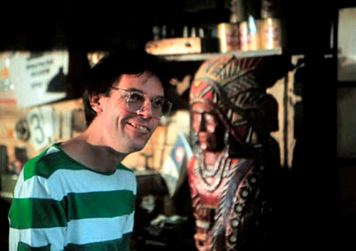 A man in a striped shirt smiles next to a statue of a Native American.