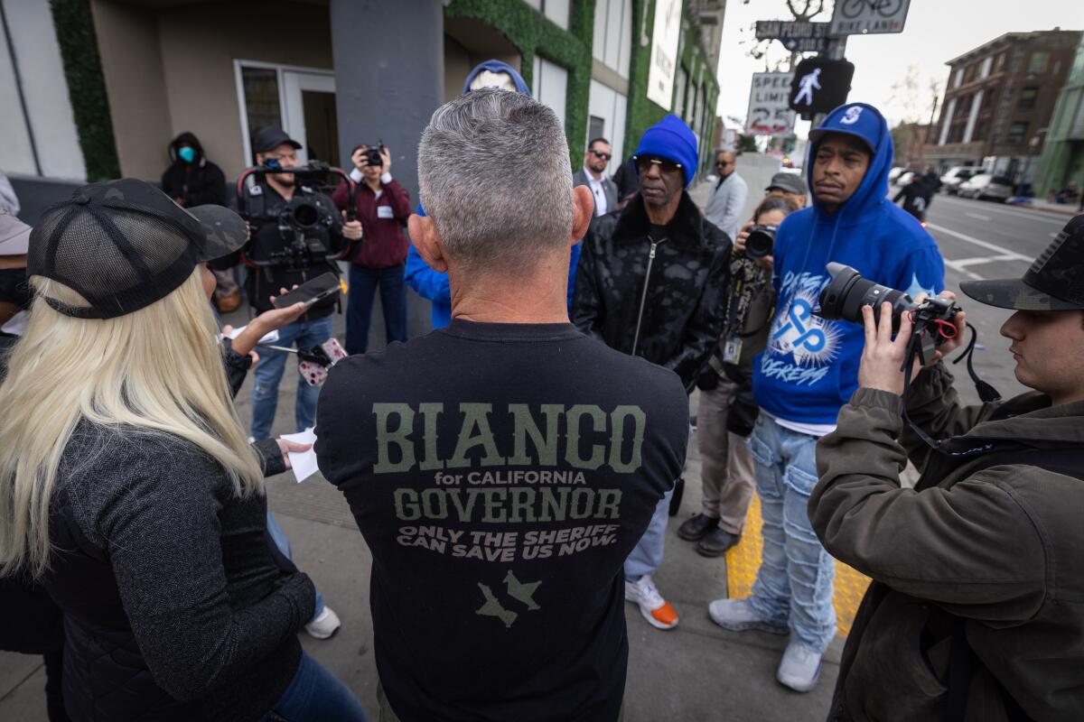 A man wearing a Bianco for Governor shirt with his back to the camera stands with people on Skid Row