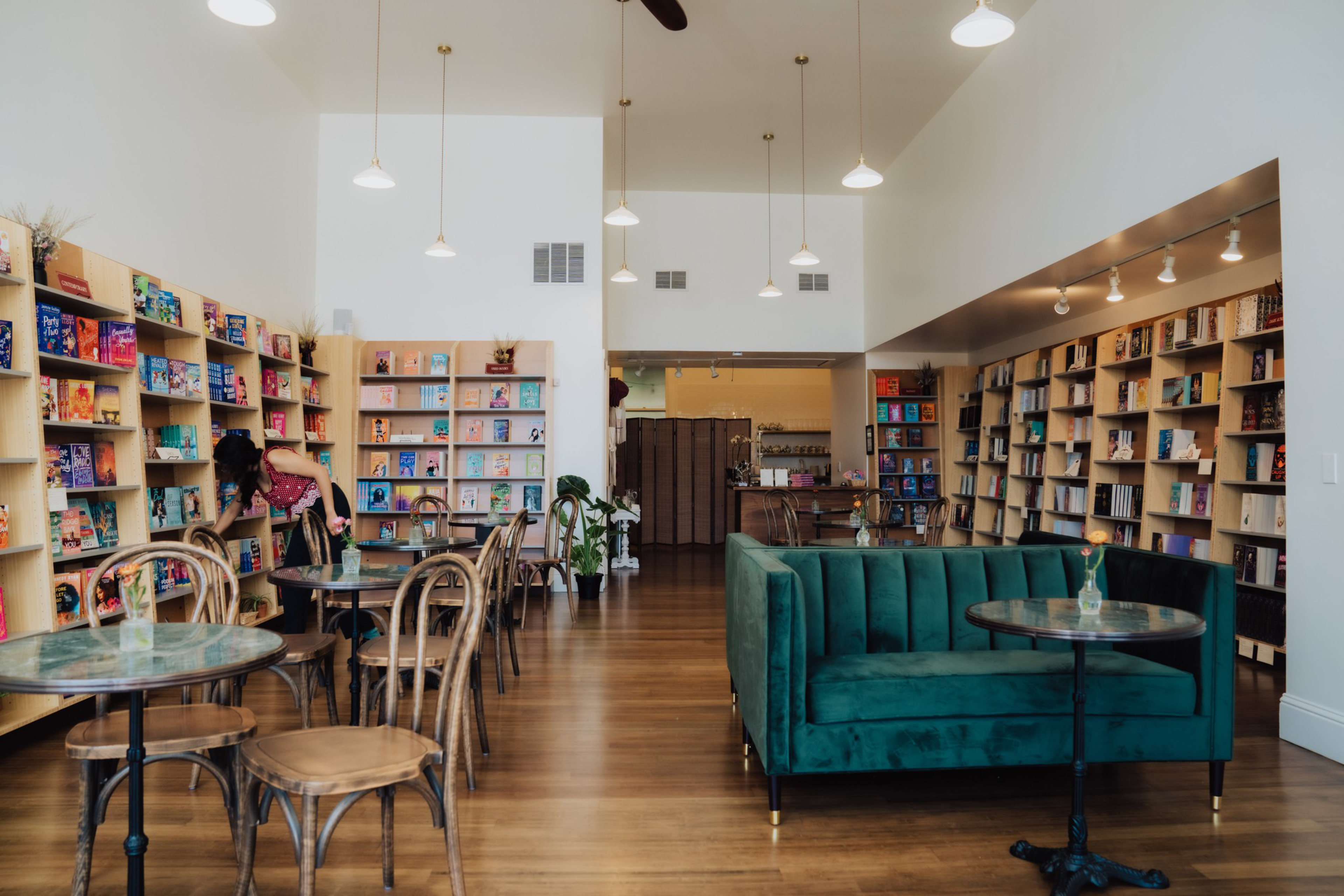 A cozy bookstore with wooden floors, round tables with chairs, a green velvet couch, and shelves filled with colorful books along the walls.