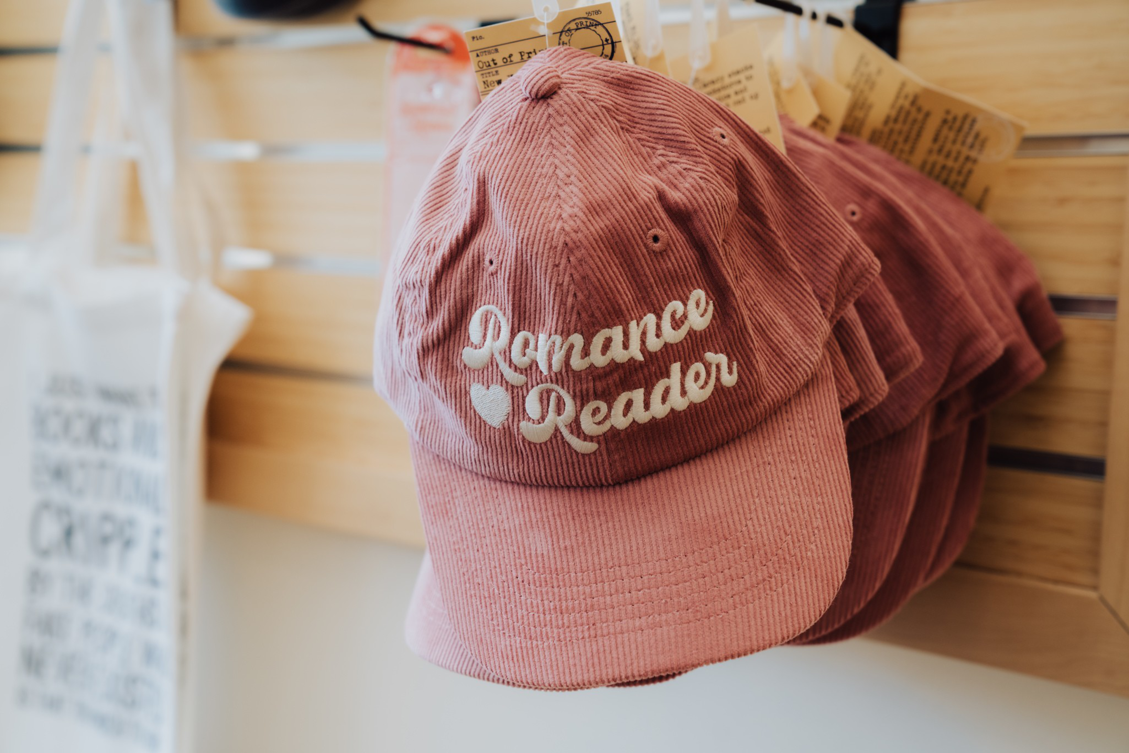 Several pink corduroy baseball caps hang on hooks, each embroidered with the phrase “Romance Reader” and a small heart.