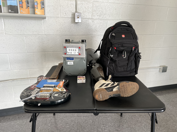 A small black table is seen in a room, there is a white brick wall behind the table and a gray linoleum floor under it. On the table an electric guitar, and electric meter, a stack of baseball cards, a prosthetic leg with an Adidas shoe attached, and a black backpack are depicted.