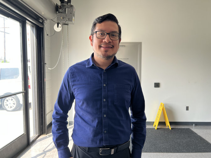 A man is seen in a white room. He wears a dark blue shirt, black glasses, a black belt and black slacks. Behind him to his left there is a black carpet with a yellow "Wet Floor" sign on it.