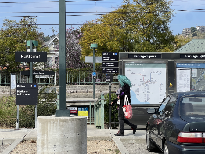 A train station is seen from its parking lot. A person with blue hair walks across a walkway. There are several signs to their right side. A Metro map that says "Heritage Square" is closest to the person. Down the walkway a sign says "Platform 1" below it there is a smaller sign that says "Metro Platform 1 to Pomona".