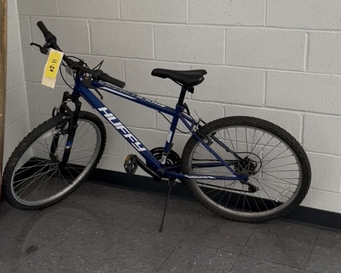 A blue "Huffy" bicycle is propped up by its kickstand next to a white brick wall and a gray tiled floor. There is a yellow tag on the handlebars.