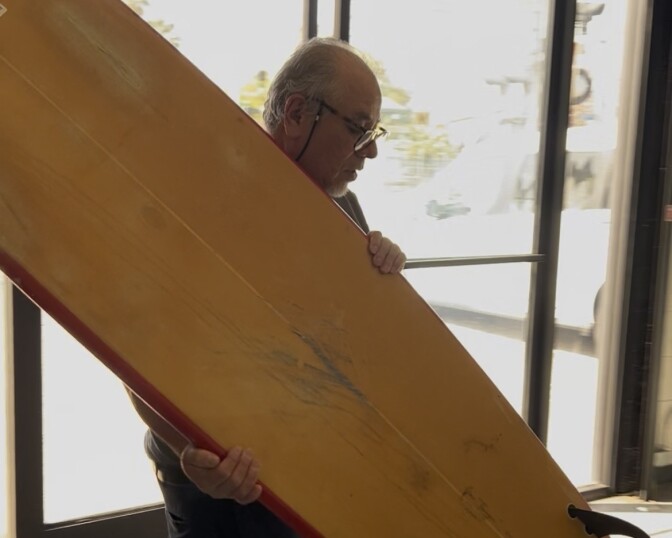 A man carries a beat up yellow surfboard. He wears chain-glasses and there are glass doors behind him where the sun is brightly filtering through.