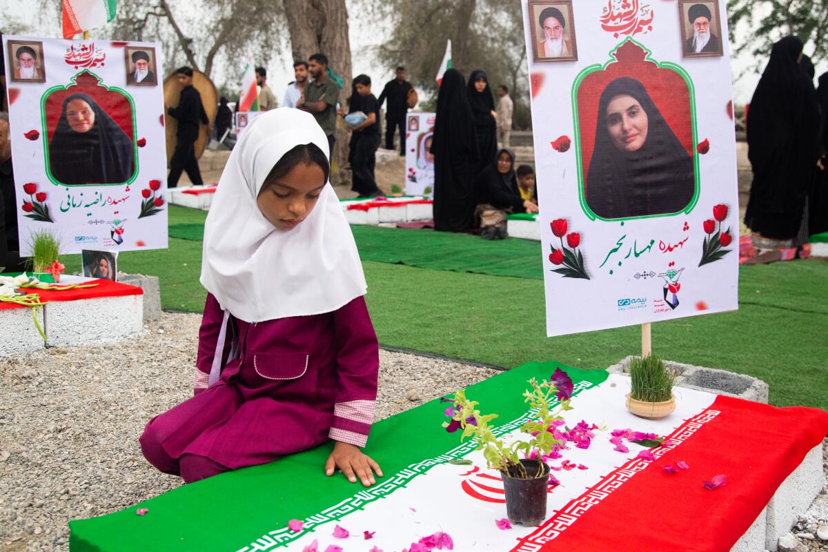A young girl in a white head covering sits next to a flag-draped gravesite.