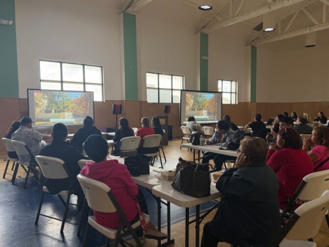 A large room with dozens of people sitting at tables looking at two screens with projectors displayed on them.