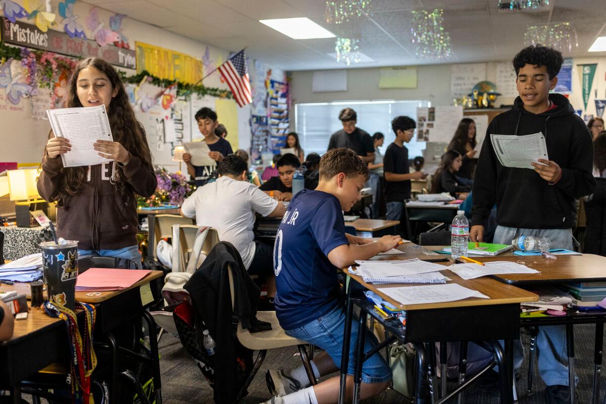 Some students read from the sheet of paper they are holding while standing, as others sit at their desks in class