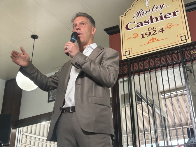 Man in grey suit holds microphone in front of sign that says "cashier"