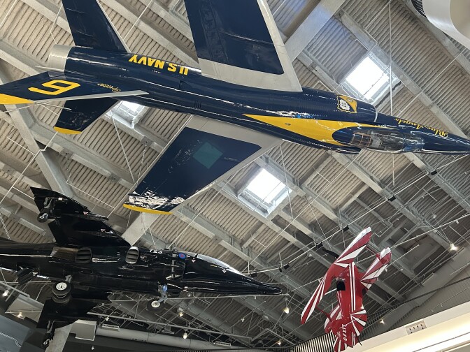 The bottom of three aircraft suspended from the ceiling on wires in a museum gallery. One aircraft is blue and yellow with the words "U.S. Navy" on the side. The aircraft below it is all black, and the aircraft to the right is red with white stripes.