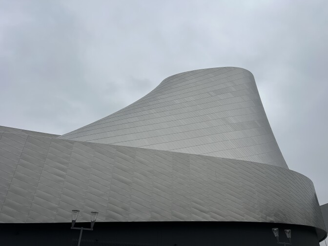 The exterior of a large silver building with a roundish structure reaching out toward the gray, overcast sky.