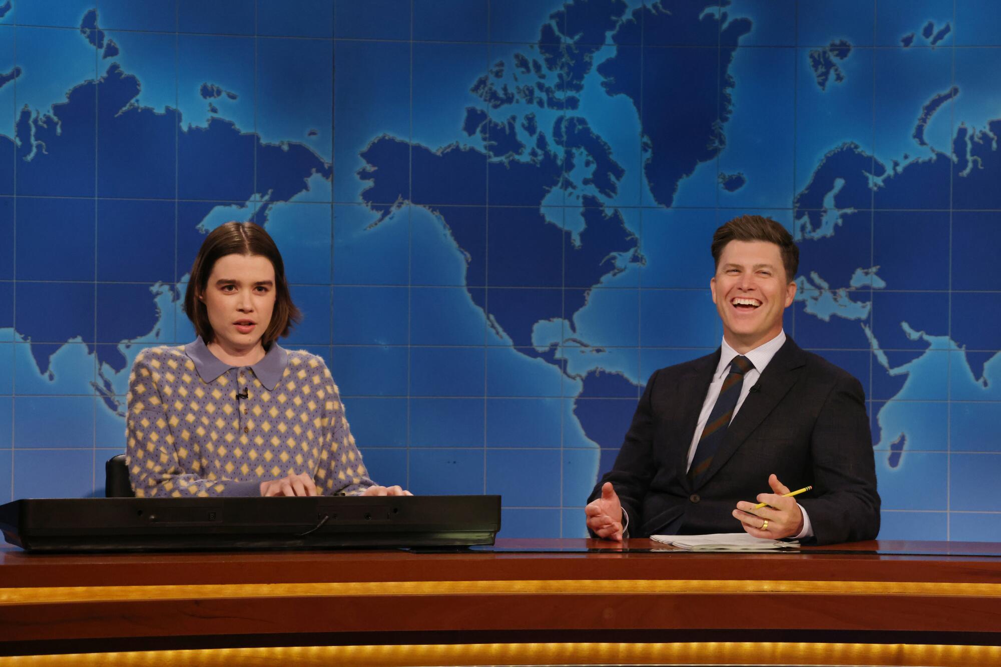 A woman plays the keyboard at a desk next to a man in suit who is laughing.