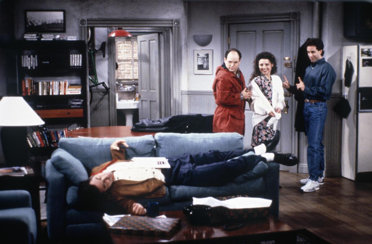 A man lies on a sofa while two other men and a woman stare at him