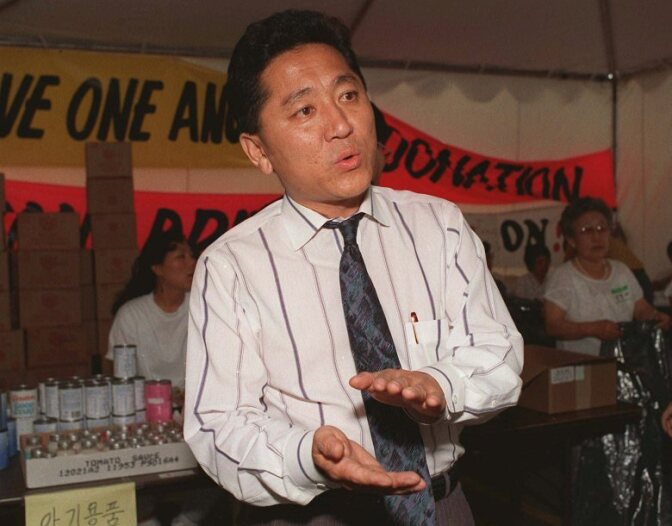 A man with medium skin tone, wearing a white striped shirt and tie, speaks and looks out of frame as he gestures with his hands. There are people behind in at tables with boxes and cans on top of them.