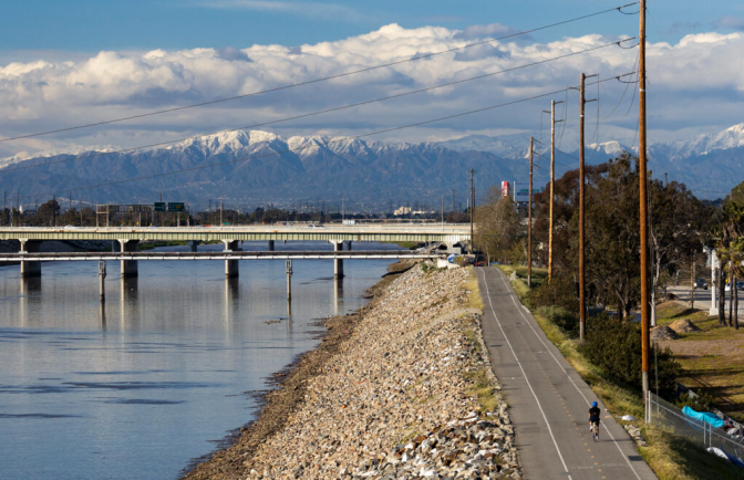A slightly high angle view of a bicylist riding along a bike path near a river towards a bridge with large mountains in the background.