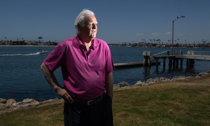 A man with white hear wearing a purple short sleeve shirt stands with his hand on his hip, looking into the distance. Behind him is a harbor.