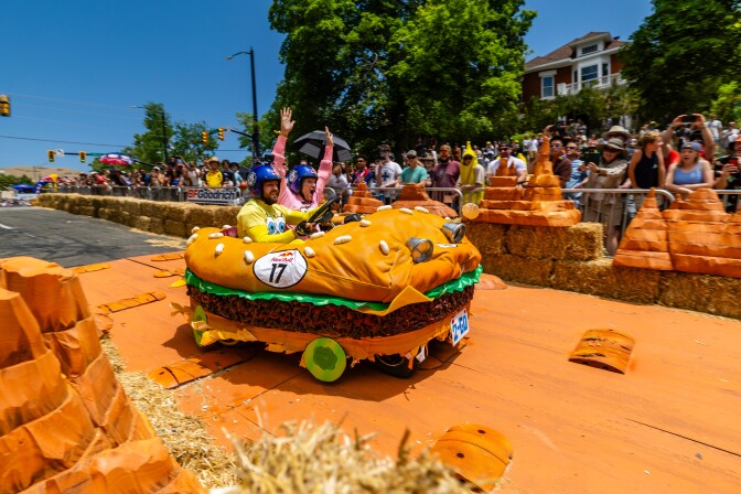 A race course lined with hay bails and orange flooring, with a soapbox designed to look like a big burger rolling down the track. Two people are driving the burger-car, with one wearing a yellow shirt that looks like the SpongeBob cartoon character and another wearing a pink shirt to resemble Patrick. The passenger wearing pink has both arms raised in the air.