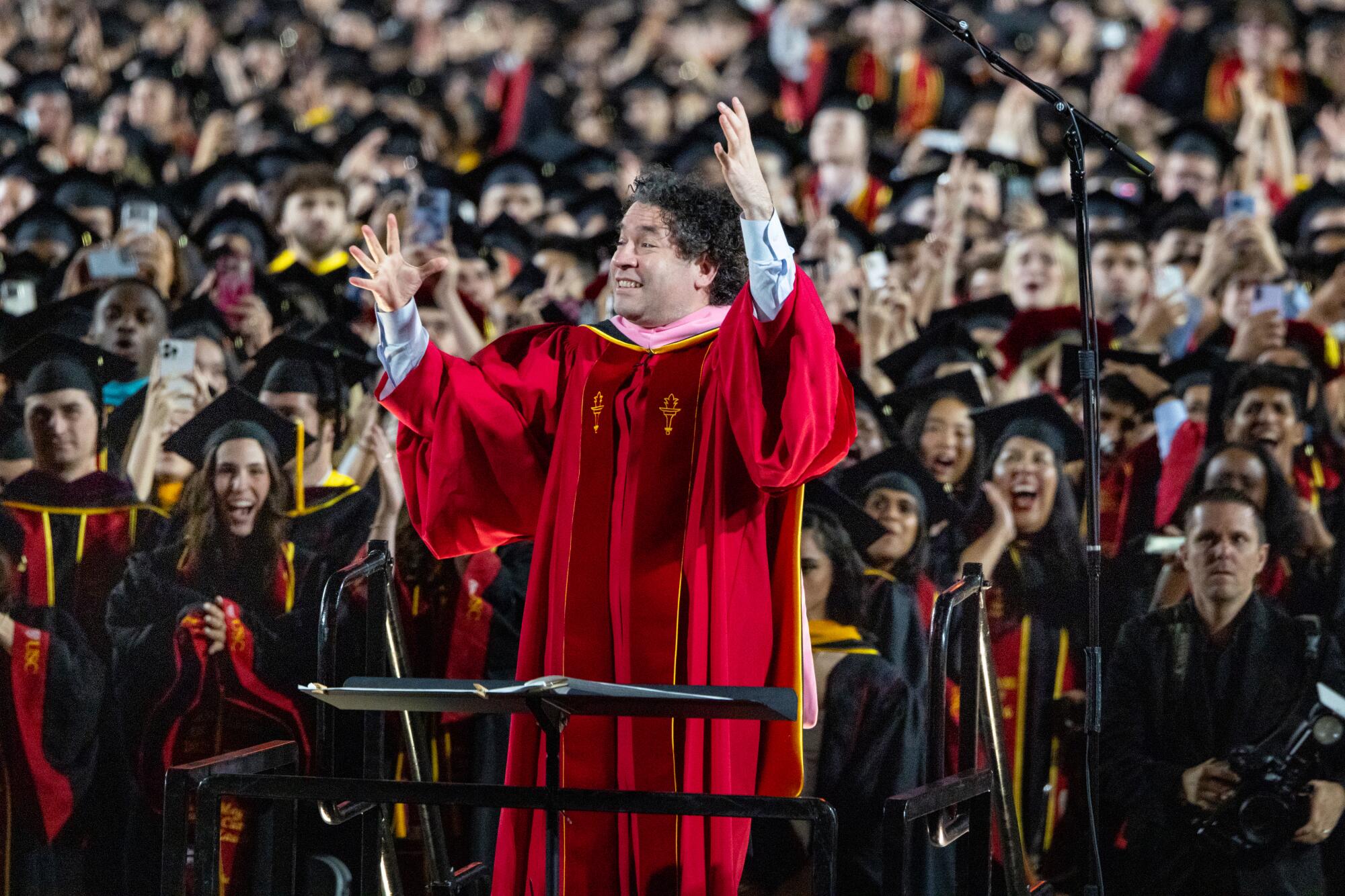 In a crimson robe, Gustavo Dudamel directs the USC band at the 2026 commencement ceremony while graduates look on.