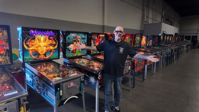 A bald man with glasses spreads his arms in front of many classic pinball machines.