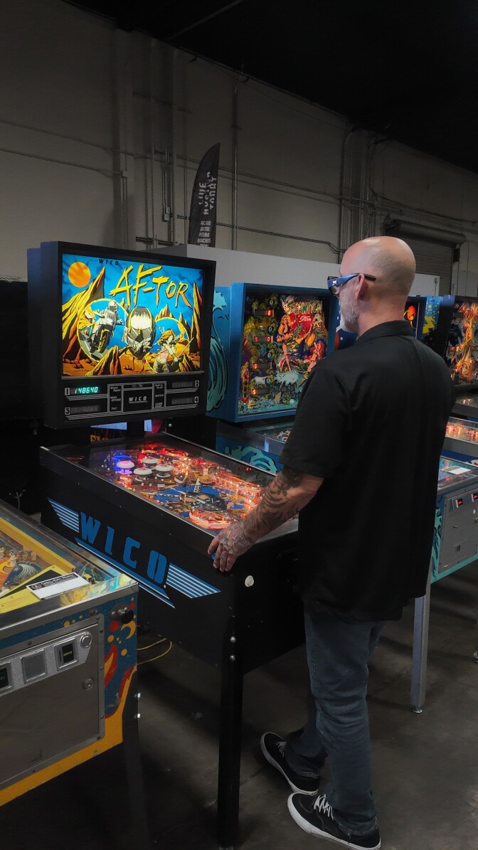 A bald man with glasses plays a pinball machine.